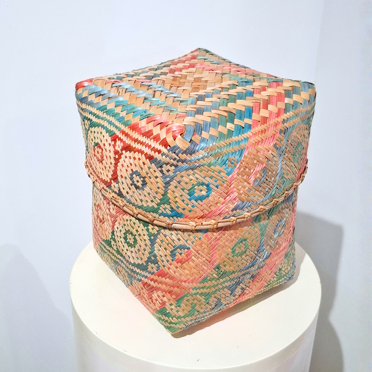 A square woven basket with a lid, featuring a colorful geometric and circular pattern in beige, pink, blue, and green, displayed on a white cylindrical pedestal against a light gray background.