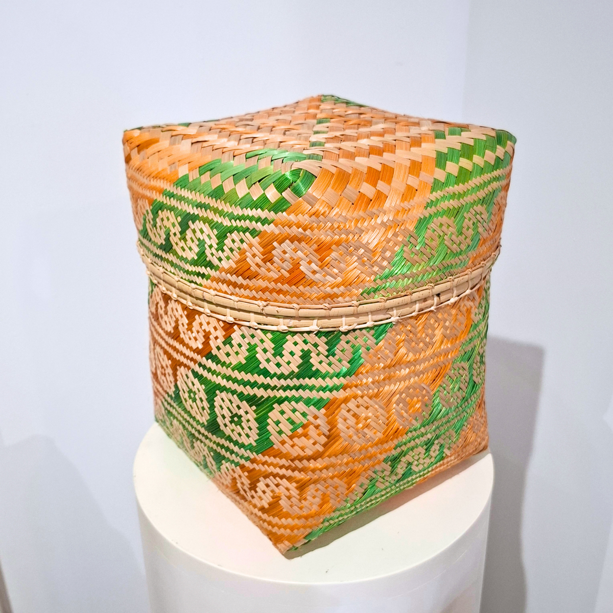 A square hand-woven basket with geometric orange, green, and beige patterns, displayed on a white cylindrical pedestal against a light gray background.