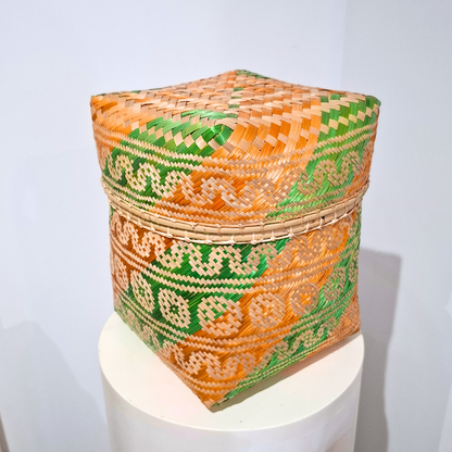 A square hand-woven basket with geometric orange, green, and beige patterns, displayed on a white cylindrical pedestal against a light gray background.