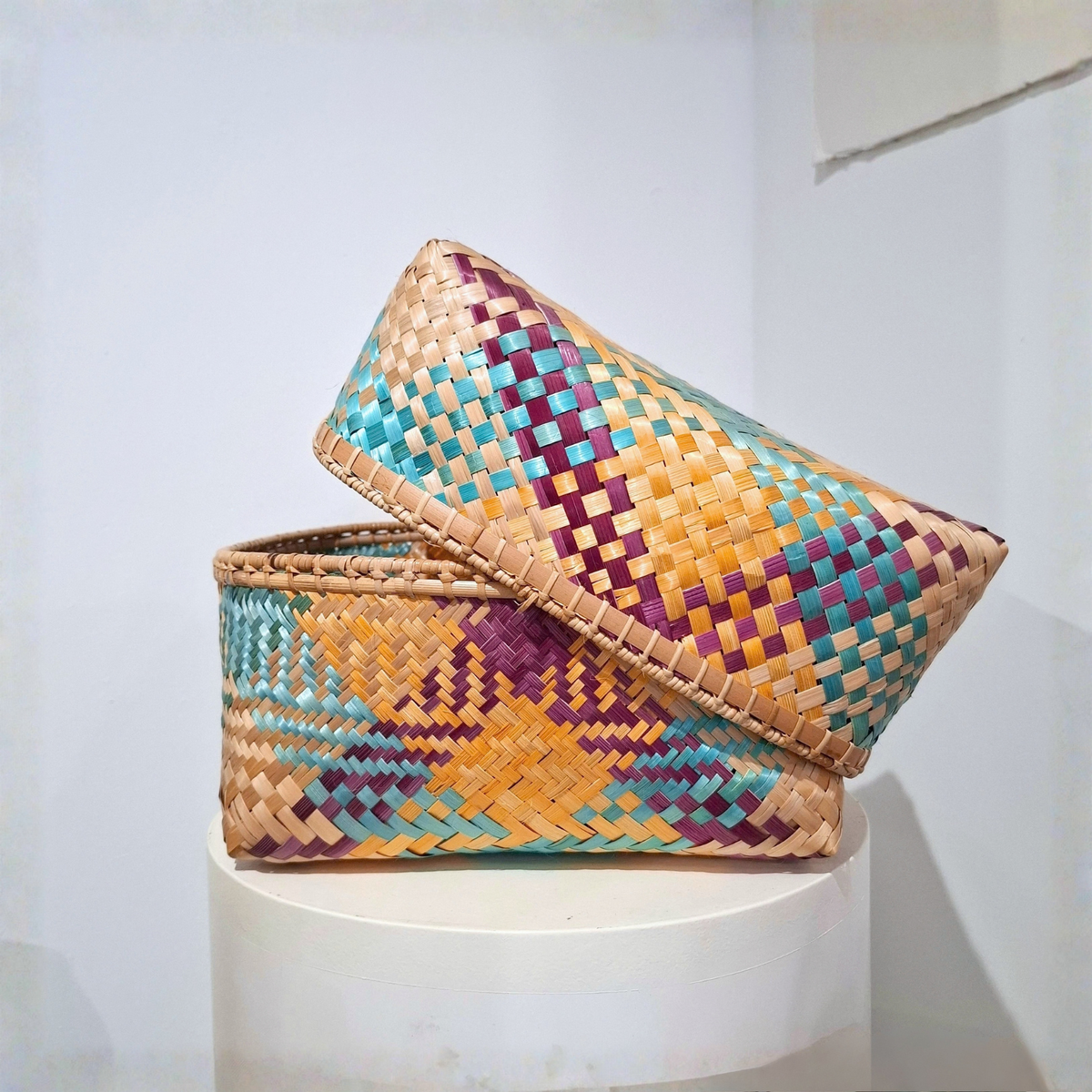 A woven rectangular basket with matching lid, displayed on a white pedestal against a plain light background. The basket features a multicolor geometric pattern with natural tan, turquoise, purple, and orange-brown woven strands.