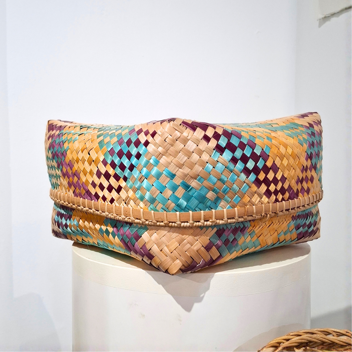 A woven rectangular basket made from natural tan strips with geometric diamond patterns in turquoise, purple, and beige, displayed on a white cylindrical pedestal against a white wall.