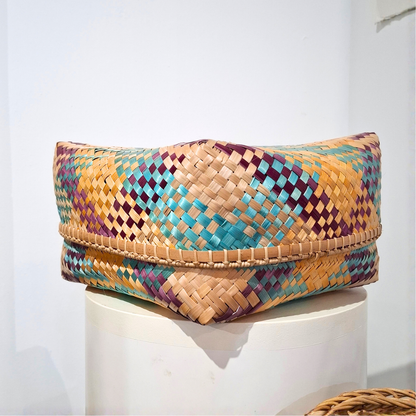 A woven rectangular basket made from natural tan strips with geometric diamond patterns in turquoise, purple, and beige, displayed on a white cylindrical pedestal against a white wall.