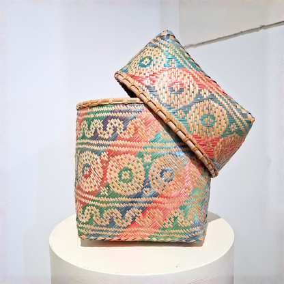 A handwoven basket with a matching lid, featuring a colorful geometric pattern in beige, red, green, and blue, displayed on a white pedestal against a white wall.