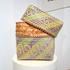 A handwoven square basket with a matching lid, displayed on a white pedestal. The basket features a natural tan woven body with a decorative patterned border in green, blue, and tan.