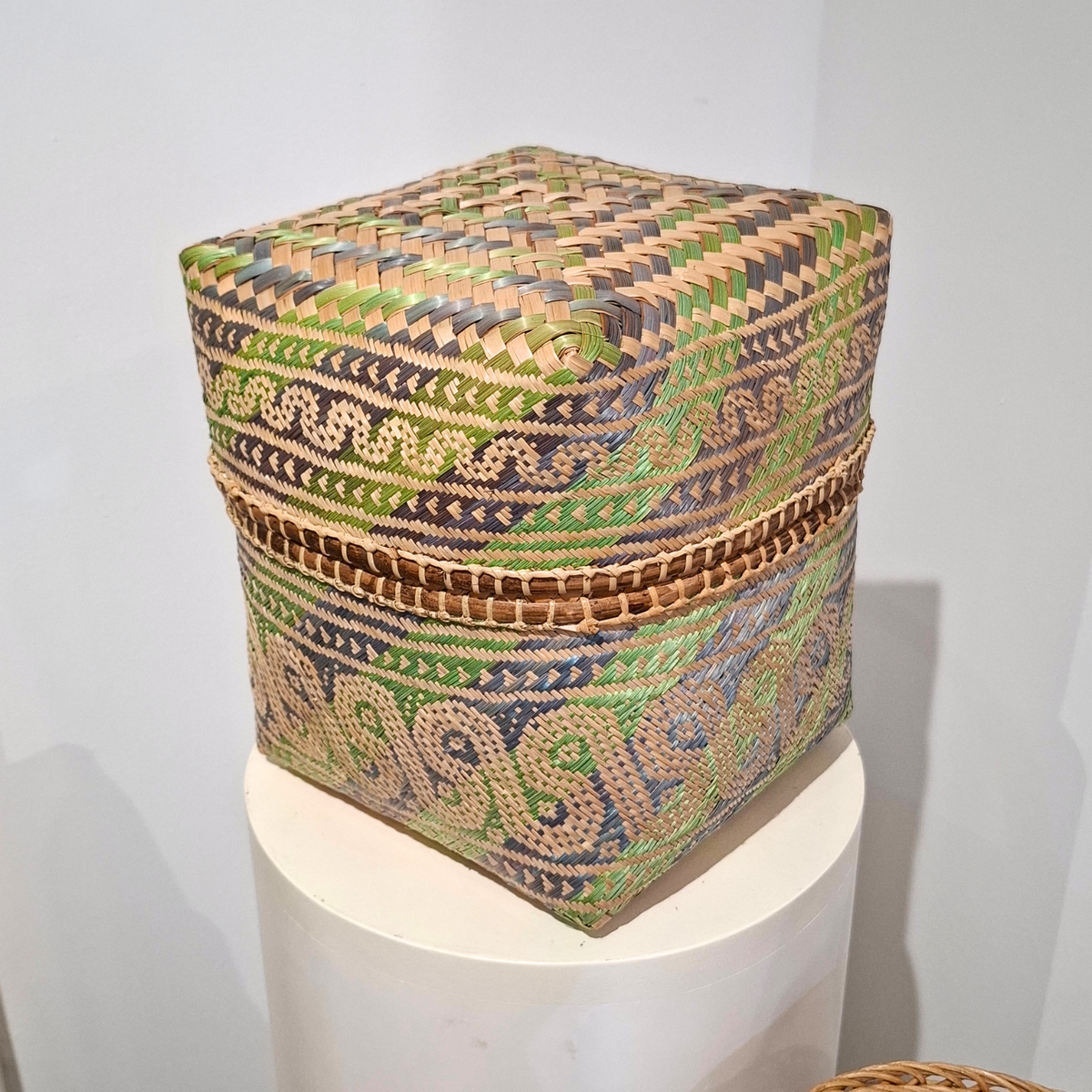 A square woven basket with a lid, featuring intricate geometric and floral patterns in green, blue, brown, and beige, displayed on a white cylindrical pedestal.