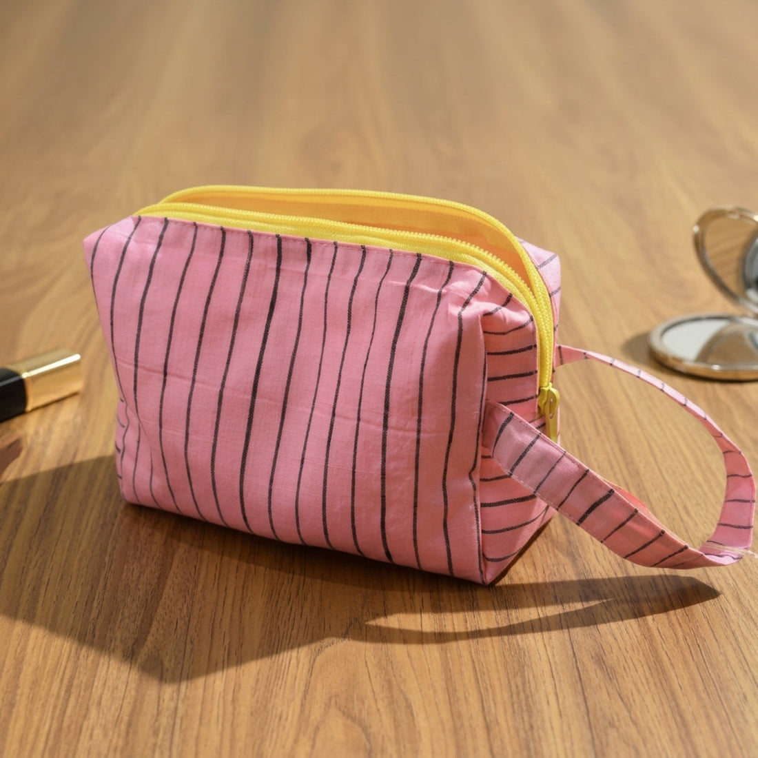 Pink striped toiletry bag with a yellow zipper and wrist strap, placed on a wooden tabletop.
