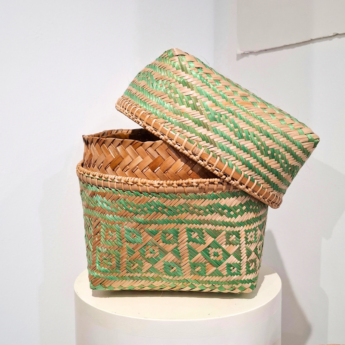 A woven square basket with a matching lid, displayed on a white pedestal against a plain light background. The basket features a geometric pattern woven from tan and green fibers.