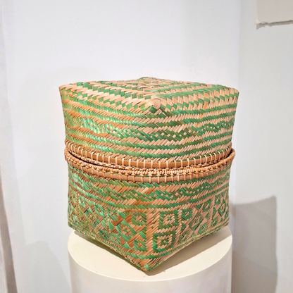 A square woven basket with a lid, featuring a pattern of green and natural tan fibers, displayed on a white pedestal against a white wall.
