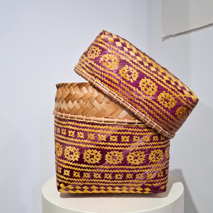 Hand-woven rectangular basket with a matching lid. The basket feature a geometric pattern in purple and yellow on a natural tan base, displayed on a white pedestal against a light gray wall.