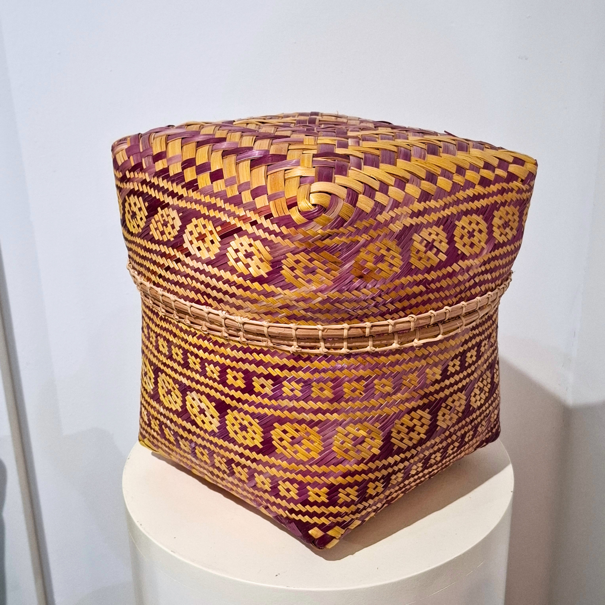 A square hand-woven basket with a lid, featuring intricate geometric patterns in purple and golden yellow, displayed on a white pedestal against a plain light background.