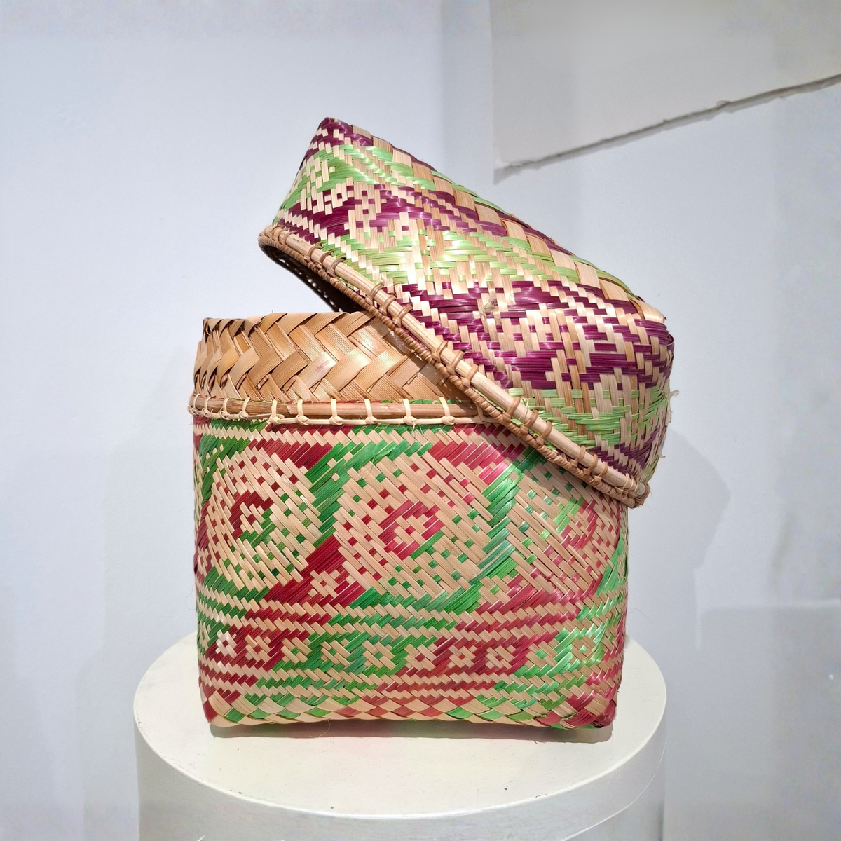 A hand-woven square basket with a matching lid, featuring intricate geometric patterns in red, green, and purple on a natural tan base, displayed on a white pedestal against a plain white wall.