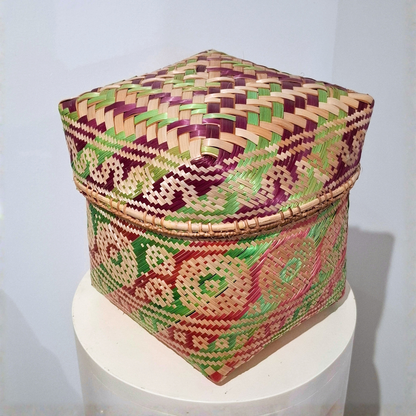 A square hand-woven basket box with a lid, featuring intricate geometric patterns in purple, green, red, and natural tan, displayed on a white cylindrical pedestal against a light gray background.